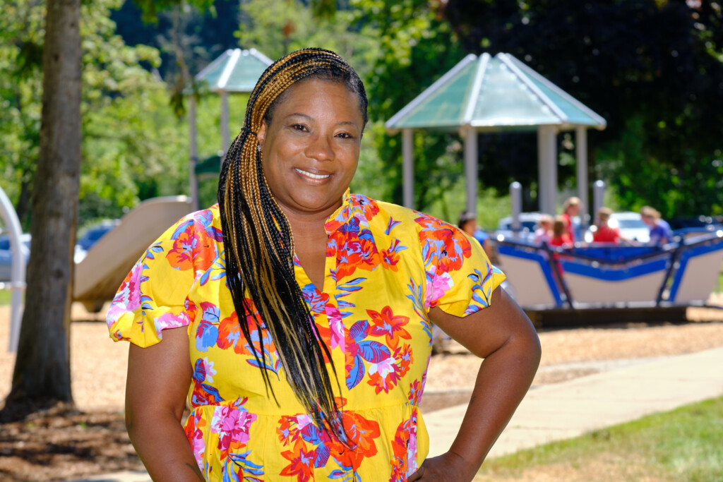 Tracye Ferguson poses in front of a playground which is blurred in the background. Tracye is wearing a bright yellow shirt with pink and blue flower designs - arms on her hips and smiling.