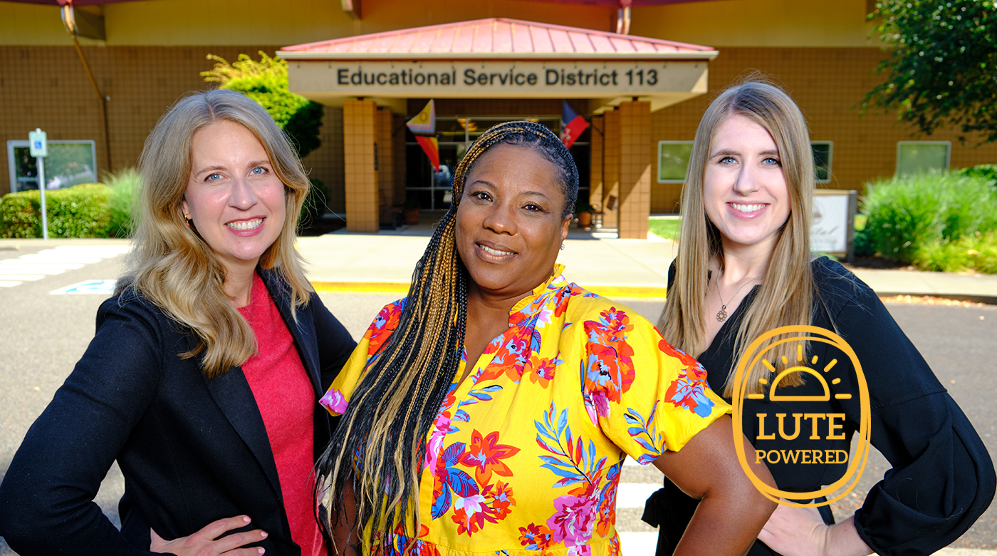 PLU alumni Kristen Jaudon ‘94 standing on the left side of the image, Tracye Ferguson ‘94 is standing in the middle of th eimage and Kate Hall ‘17 stands in the right of the images and poses side by side in a group photo in front the Educational Service District 113 building. The image displays a sunny and bright day with all three subjects smiling at the camera.