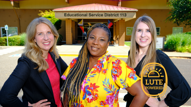 PLU alumni Kristen Jaudon ‘94 standing on the left side of the image, Tracye Ferguson ‘94 is standing in the middle of th eimage and Kate Hall ‘17 stands in the right of the images and poses side by side in a group photo in front the Educational Service District 113 building. The image displays a sunny and bright day with all three subjects smiling at the camera.