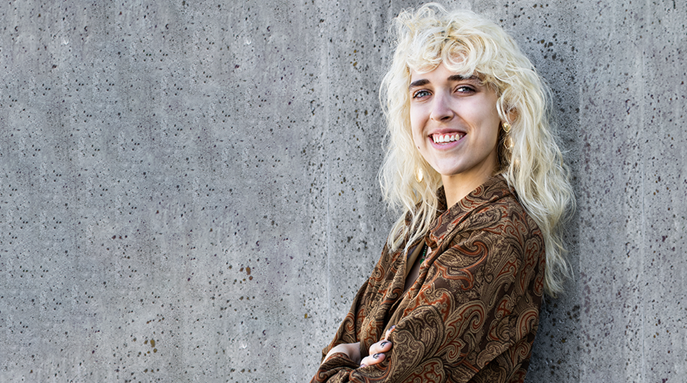 Peyton Noreen is leaning against a concrete wall with arms folded, a bright smile, and wearing a brown button up shirt with a paisley like pattern.