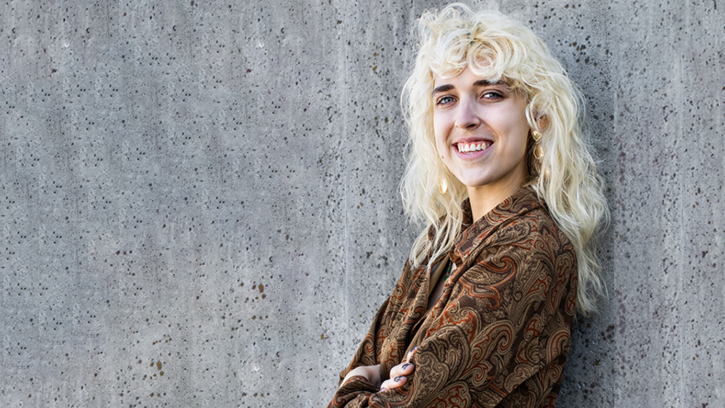 Peyton Noreen is leaning against a concrete wall with arms folded, a bright smile, and wearing a brown button up shirt with a paisley like pattern.