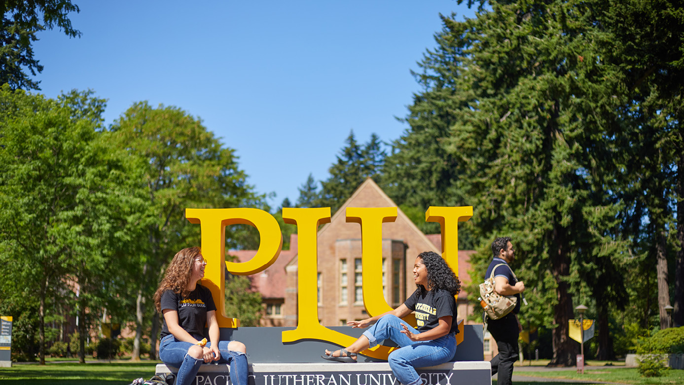 Two PLU students talk and laugh while sitting on the big gold PLU sign on the edge of campus.