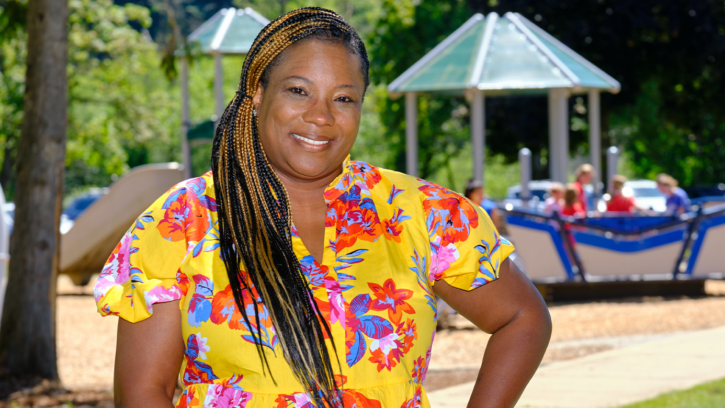 Tracye Ferguson poses in front of a playground which is blurred in the background. Tracye is wearing a bright yellow shirt with pink and blue flower designs - arms on her hips and smiling.