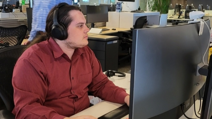 A man sitting at a desk staring at a computer monitor with over the ear headphones on in an office setting.