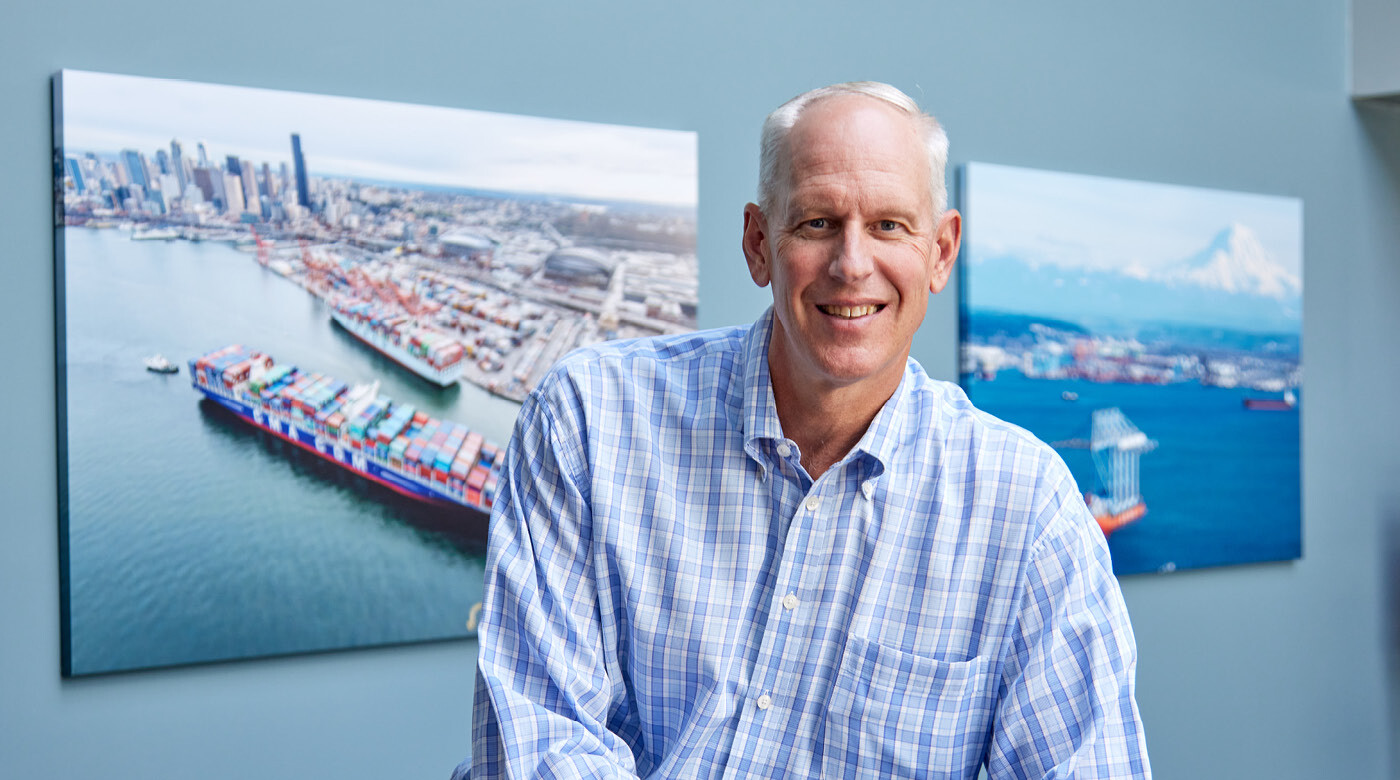 A portrait of Mark Miller in front of large photographs of the Port of Tacoma. Miller is smiling wearing a patterned buttoned up shirt. (Photo by John Froschauer/PLU)