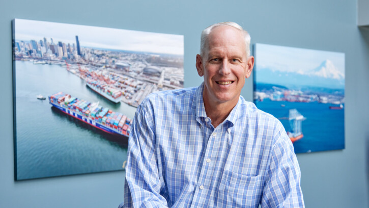 A portrait of Mark Miller in front of large photographs of the Port of Tacoma. Miller is smiling wearing a patterned buttoned up shirt. (Photo by John Froschauer/PLU)