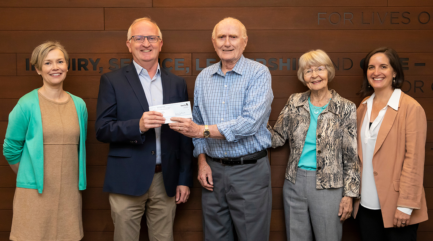 A group of men and women posing and smiling in front of a brown wall. There are five subjects in the photo and starting from left to right: A women in a turquoise cardigan over a brown dress, next to the woman is a man standing in khakis, a blue jacket holding a paper check along with another gentlleman holding the same check. Next to the men to theright are two women standing side by side with one another.