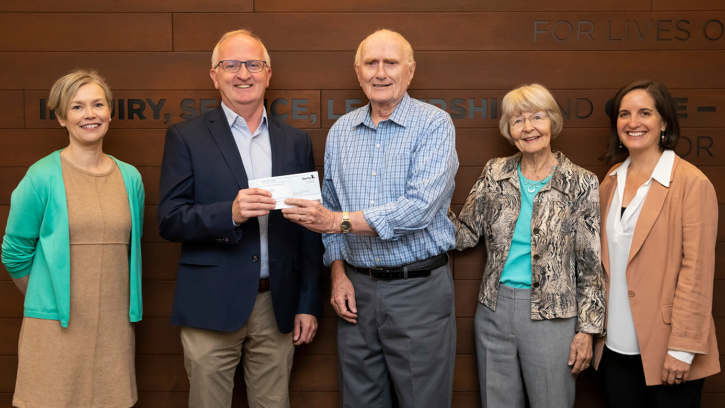 A group of men and women posing and smiling in front of a brown wall. There are five subjects in the photo and starting from left to right: A women in a turquoise cardigan over a brown dress, next to the woman is a man standing in khakis, a blue jacket holding a paper check along with another gentlleman holding the same check. Next to the men to theright are two women standing side by side with one another.