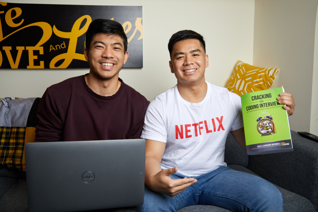 Cody Uehara ’22 (left) and Adrian Ronquillo '22 (right) sitting on a couch and smiling. Adrian is holding a book with a green cover and Cody is behind a laptop placed on the coffee table.