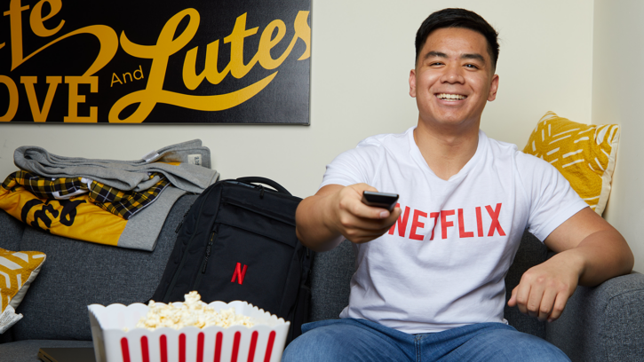 Adrian Ronquillo is sitting on a couch with a big smile on his face, wearing a white t-shirt with a red Netflix logo and pointing a remote at us as if we were the television. A box of popcorn can be seen partially covering a Netflix backpack that is placed on the couch next to Adrian in the foreground to the left of the image. A sign on the background wall reads, "Live, Love, and Lutes."