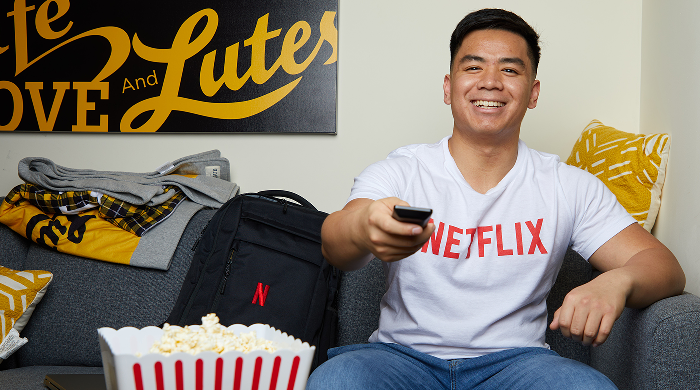 Adrian Ronquillo is sitting on a couch with a big smile on his face, wearing a white t-shirt with a red Netflix logo and pointing a remote at us as if we were the television. A box of popcorn can be seen partially covering a Netflix backpack that is placed on the couch next to Adrian in the foreground to the left of the image. A sign on the background wall reads, "Live, Love, and Lutes."