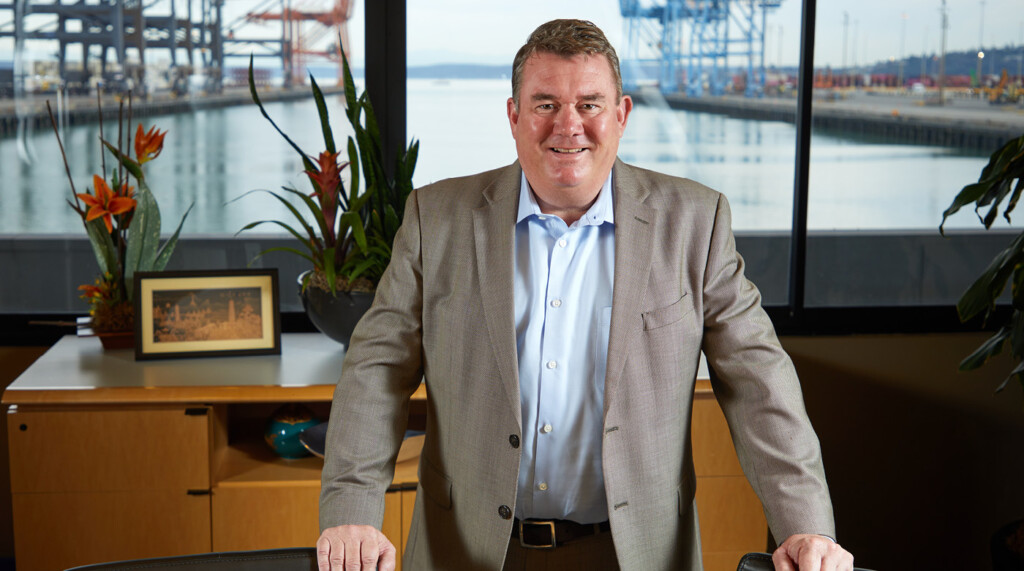 Eric Johnson ’83 standing in a Port of Tacoma meeting room. His hands rest on the backs of two chairs and we can see the port behind him through giant windows. Eric is wearing a grey blazer, blue shirt and is smiling confidently.