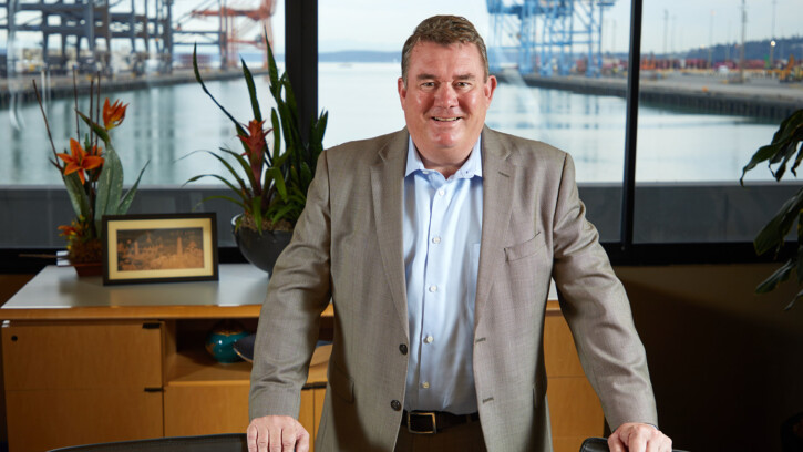 Eric Johnson ’83 standing in a Port of Tacoma meeting room. His hands rest on the backs of two chairs and we can see the port behind him through giant windows. Eric is wearing a grey blazer, blue shirt and is smiling confidently.