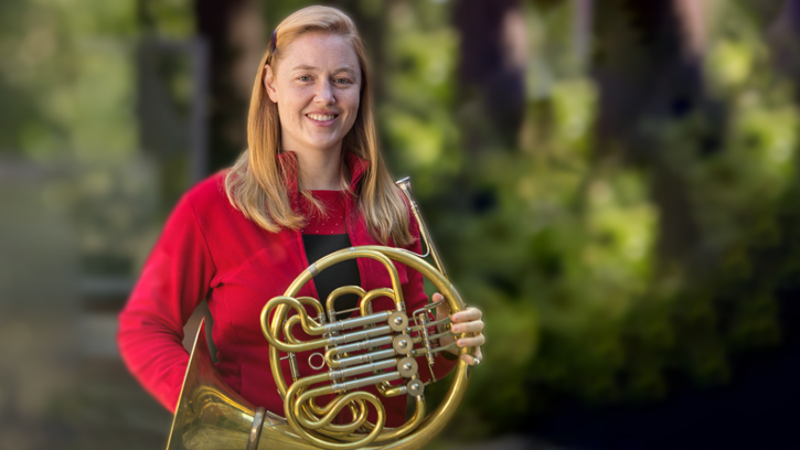 Gina Gillie poses for a photograph while holding a French horn and wearing a red zip-up sweater against a blurry background of natural green foliage.