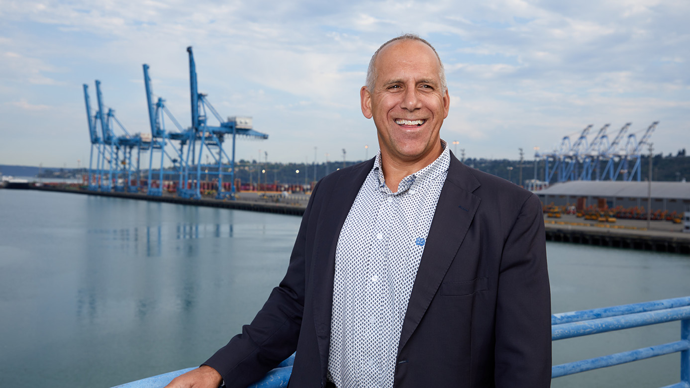 John Wolfe wearing a dress shirt and blazer. He is smiling with his hand on a metal rail and the Port of Tacoma behind him.