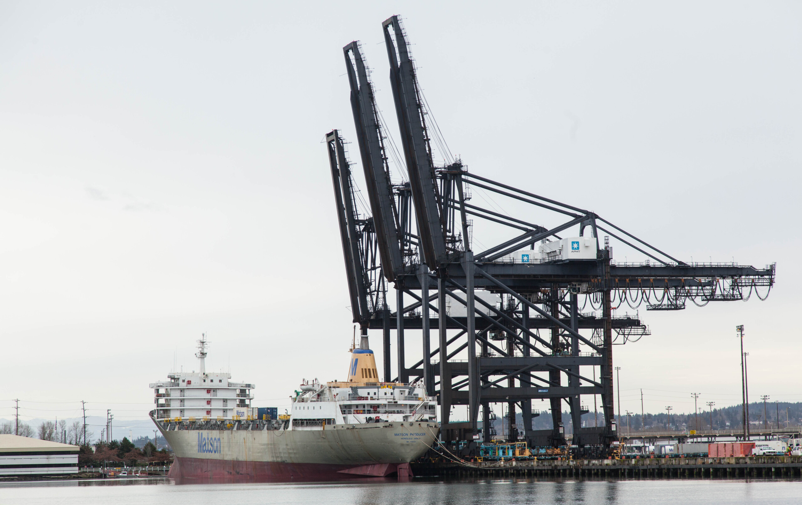 A further away photo of a boat docked in the Port of Tacoma. Large crane/convey belts rise above and next to the boat.