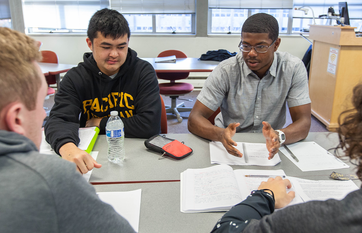 Two male students are facing the camera and discussing topics in a classroom with two other students with the back of their heads visible in the image.