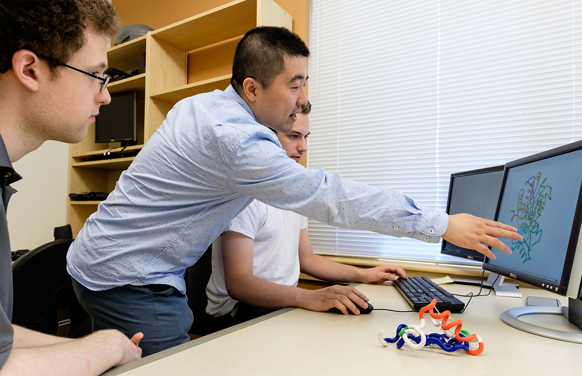 A professor pointing to a computer monitor while two students look on.