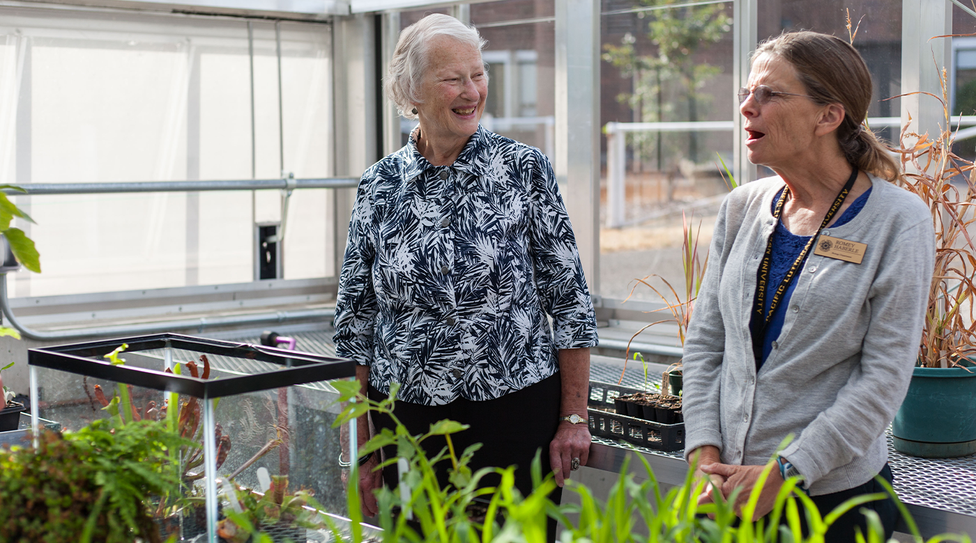 Carol Sheffels Quigg (on the left) smiles at Romey Haberle (on the right), Associate Professor of Biology, in PLU's greenhouse. As part of the foreground, blurred green plants can be seen in the bottom right corner.