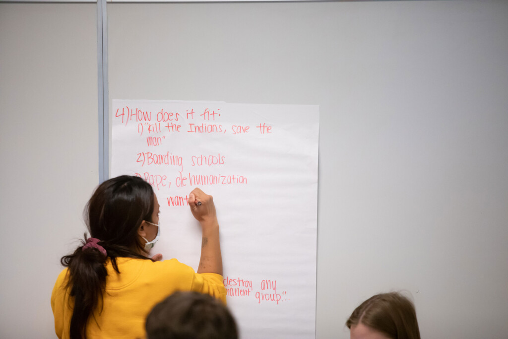 Students write presentation notes on a large white piece of paper that is taped to a wall.