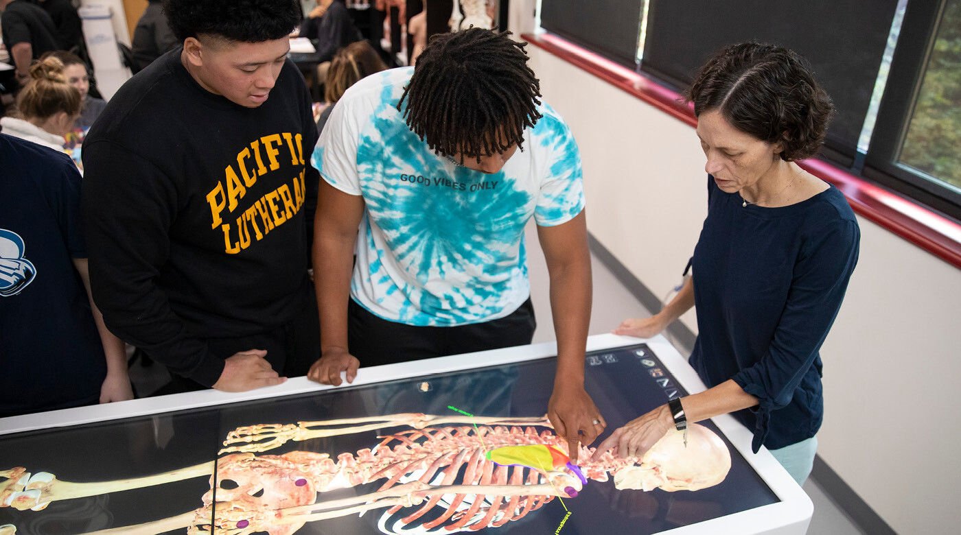 Professor Michelle Crites and two students stand around the virtual dissection table. The table's large screen displays a lifesize human skeleton system.