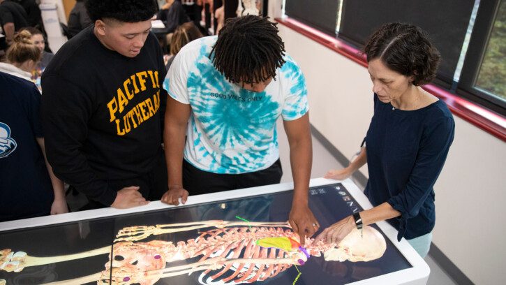 Professor Michelle Crites and two students stand around the virtual dissection table. The table's large screen displays a lifesize human skeleton system.