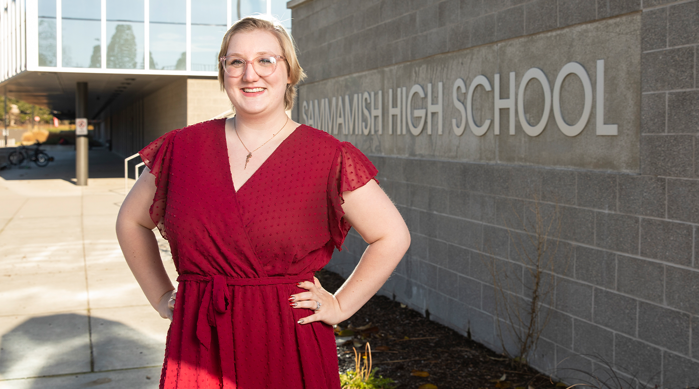 PLU alumna Becca Anderson ‘19, ‘22 is posing in front of Sammamish High School in a red dress with both hands on her hips and looking at the camera with a warm smile.