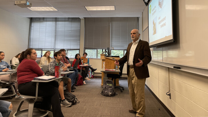 Holocaust survivor Peter Metzelaar stands in front of a classroom giving a presentation to about a dozen PLU students who are seated at small desks.