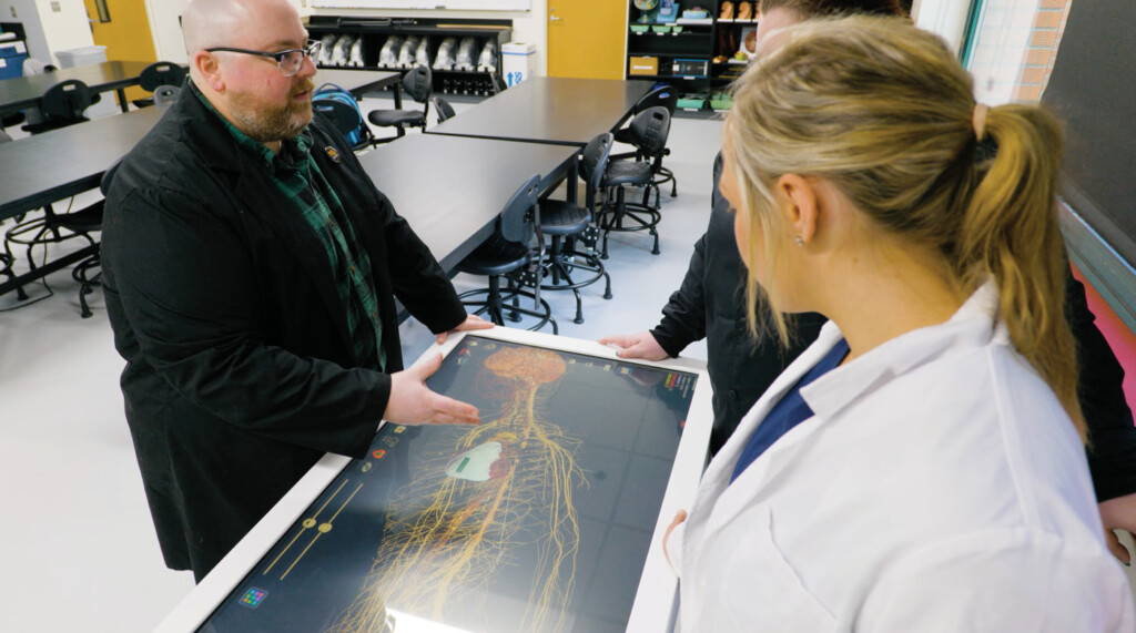 Resident Assistant Professor of Biology Jeremy Reimers speaks with two students. The three are standing around the virtual dissetion table in the A&P lab.