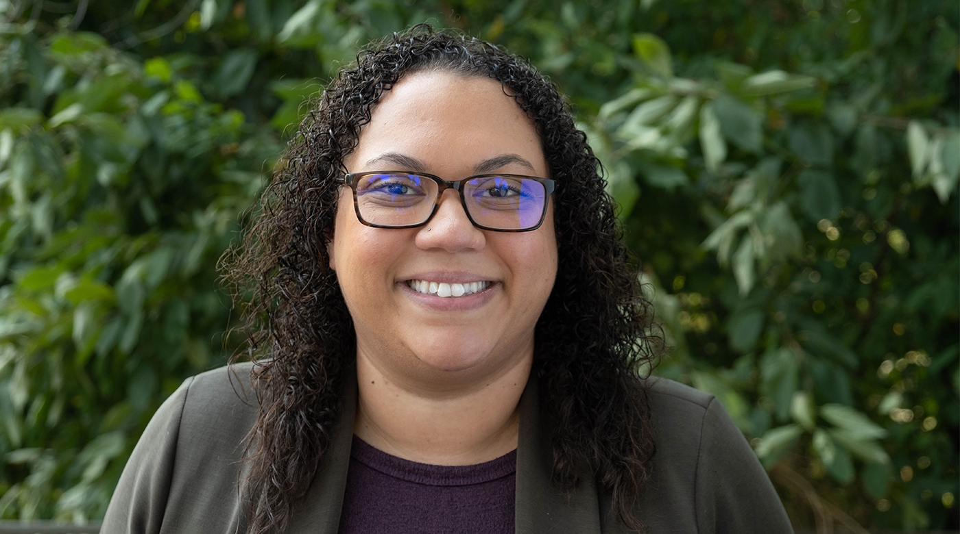 Brandi Hilliard poses in front of a greenery background that could be a bush or a tree with green leaves. Brandi is smiling and wearing a grey blazer with a purple tee in a headshot style photo.