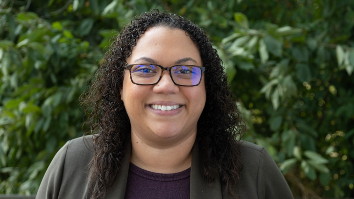 Brandi Hilliard poses in front of a greenery background that could be a bush or a tree with green leaves. Brandi is smiling and wearing a grey blazer with a purple tee in a headshot style photo.