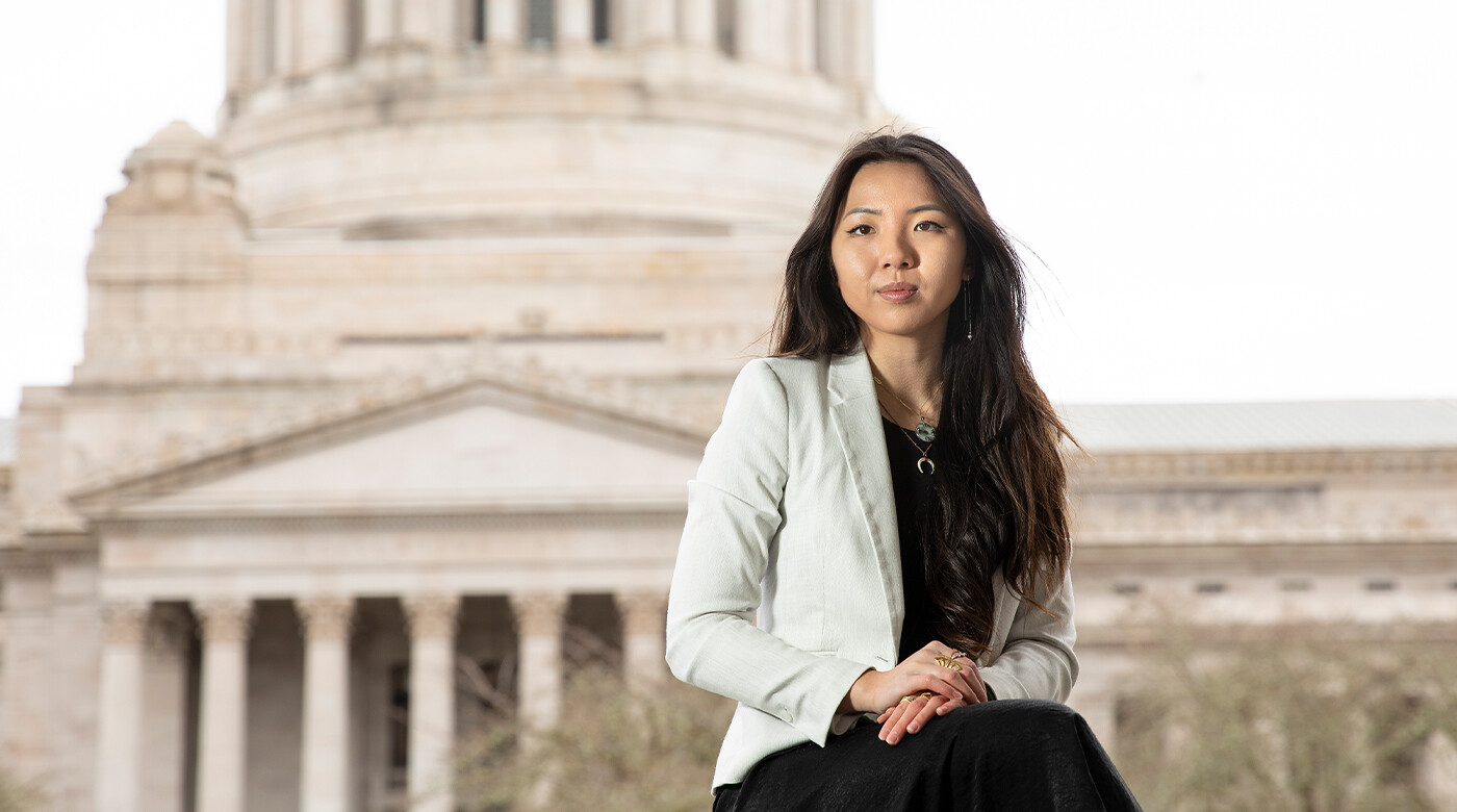 PLU student Quan Huynh ‘25 in a medium to wide portrait, posing in front of the Washington State capitol building. Huynh is sitting, arms crossed, and smiling.