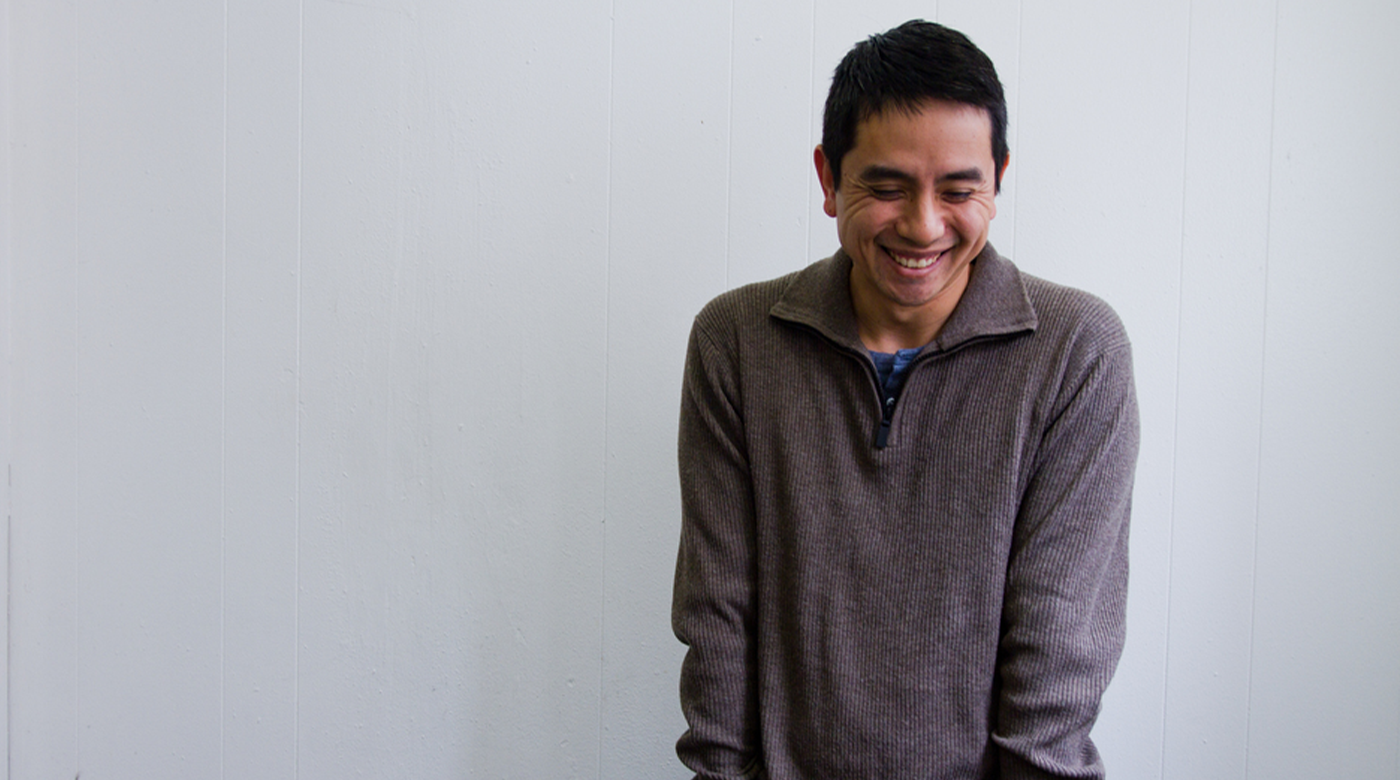 Author Minh Lê stands in front of an off white wall and looks down with a grin with both hands in his denim pants pocket.