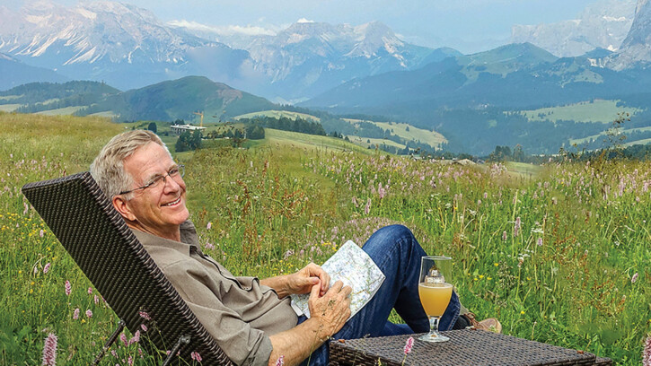 Rick Steves reclines in a chair with a grassy field and mountains behind him.