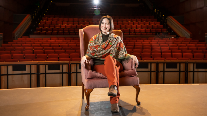 Amanda Sweger is sitting in a chair that is on a stage in a performance hall with her legs crossed. Auditorium row of seats serves as the backdrop with a spot light illuminating in the background.