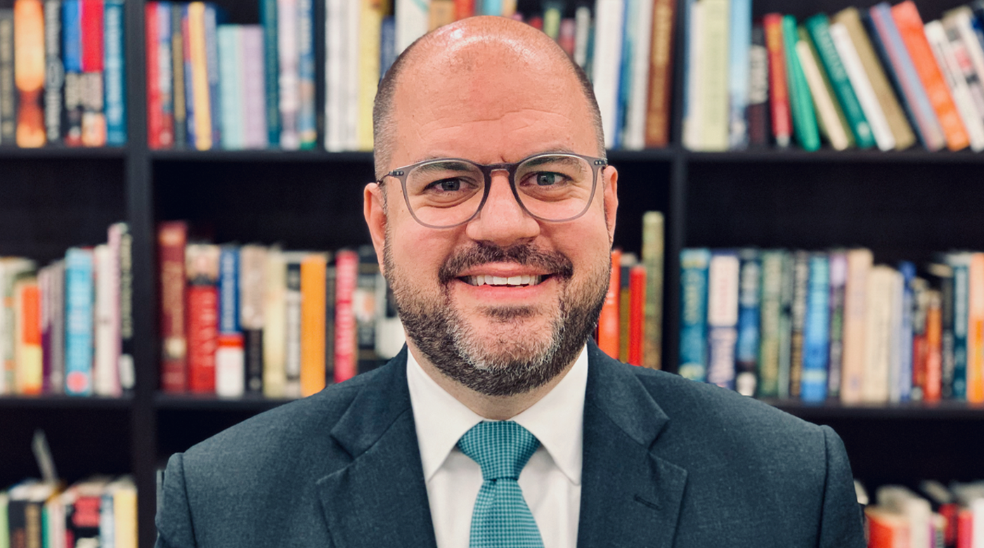 A professional portrait of Ryan Gliha in front of a shelf of books in a library.