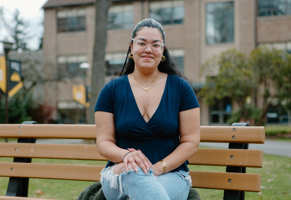 Stephanie Aparicio Zambrano sits on a brown bench on PLU's upper campus. A beautiful campus scene of grass, trees and an academic building is behind her.