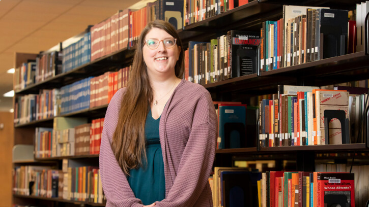 Lauren Loftis standing in a library isle filled with books. She is smiling and wearing a purple cardigan.