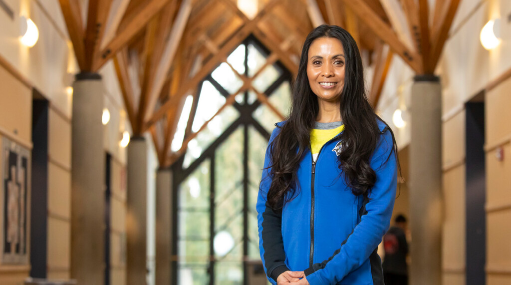 Melanie Helle smiles for a portrait while standing in a beautiful lobby space at Chief Leschi Schools. She has long dark hair and is wearing a bright blue fleece jacket.