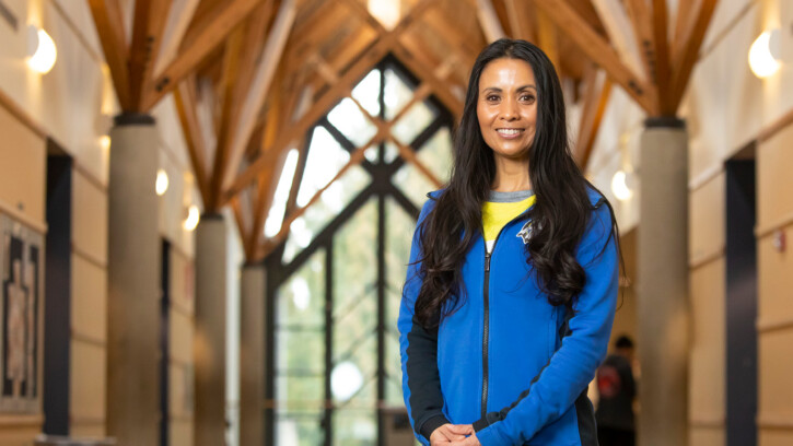 Melanie Helle smiles for a portrait while standing in a beautiful lobby space at Chief Leschi Schools. She has long dark hair and is wearing a bright blue fleece jacket.