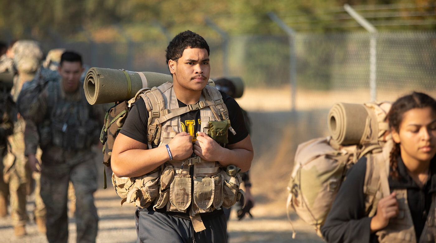A PLU student wears a black "PLU" t-shirt under his military-issue training vest and packpack. His face is stoic and focused.