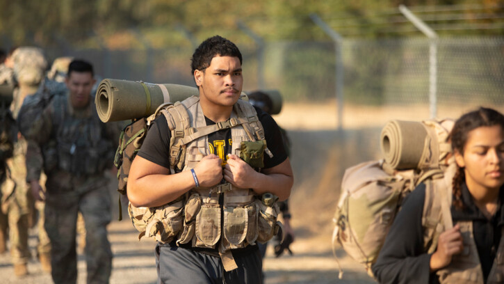 A PLU student wears a black "PLU" t-shirt under his military-issue training vest and packpack. His face is stoic and focused.