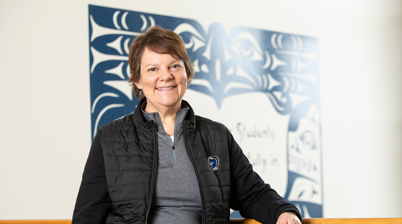 Nancy Nelson leans on bannister in the main lobby of Chief Leschi Schools. There is a large navy blue tribal mural on the wall behind her. she is smiling and wearing a black jacket with the school logo on it.