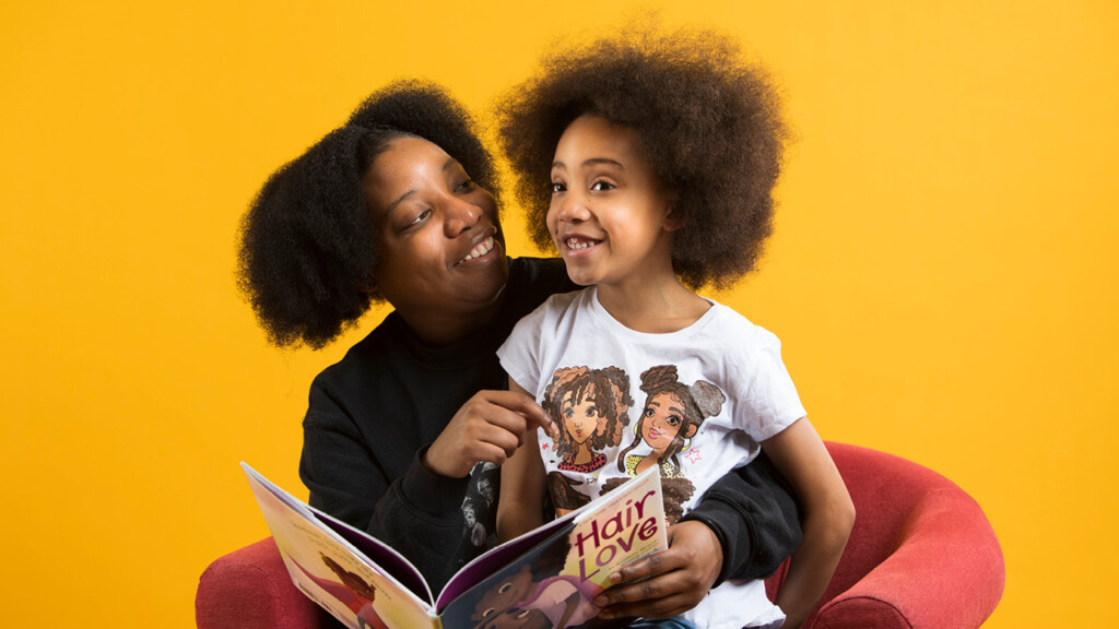 Teranejah Lucas holds her 6 year old daughter, Alanna, in her lap and reads a book to her titled 'hair love.' A gold background is behind them.