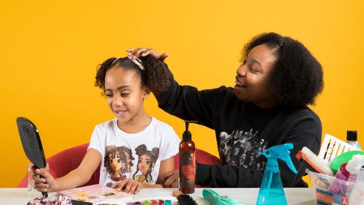 Teranejah Lucas runs her hand through her her daughter, Alanna's, hair. Alanna is holding a mirror and making a silly face. A gold background is behind them.