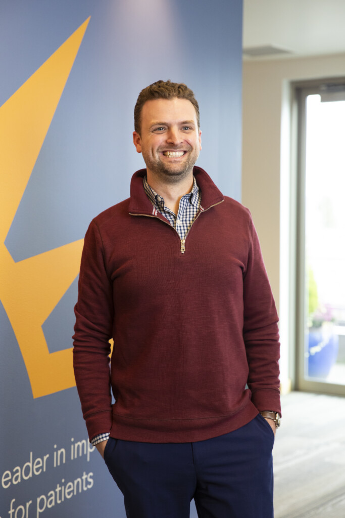 20230216_SB_Sound-Physicians_144 Michael Burris smiles while standing in the lobby of his office. There is a blue and yellow wall behind him and he is wearing a red sweater.