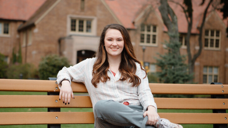 Kara Atkinson sits on a bench on PLU's upper campus with Xavier Hall behind her. She is smiling and has one of her arms wrapped around the drop of the bench.