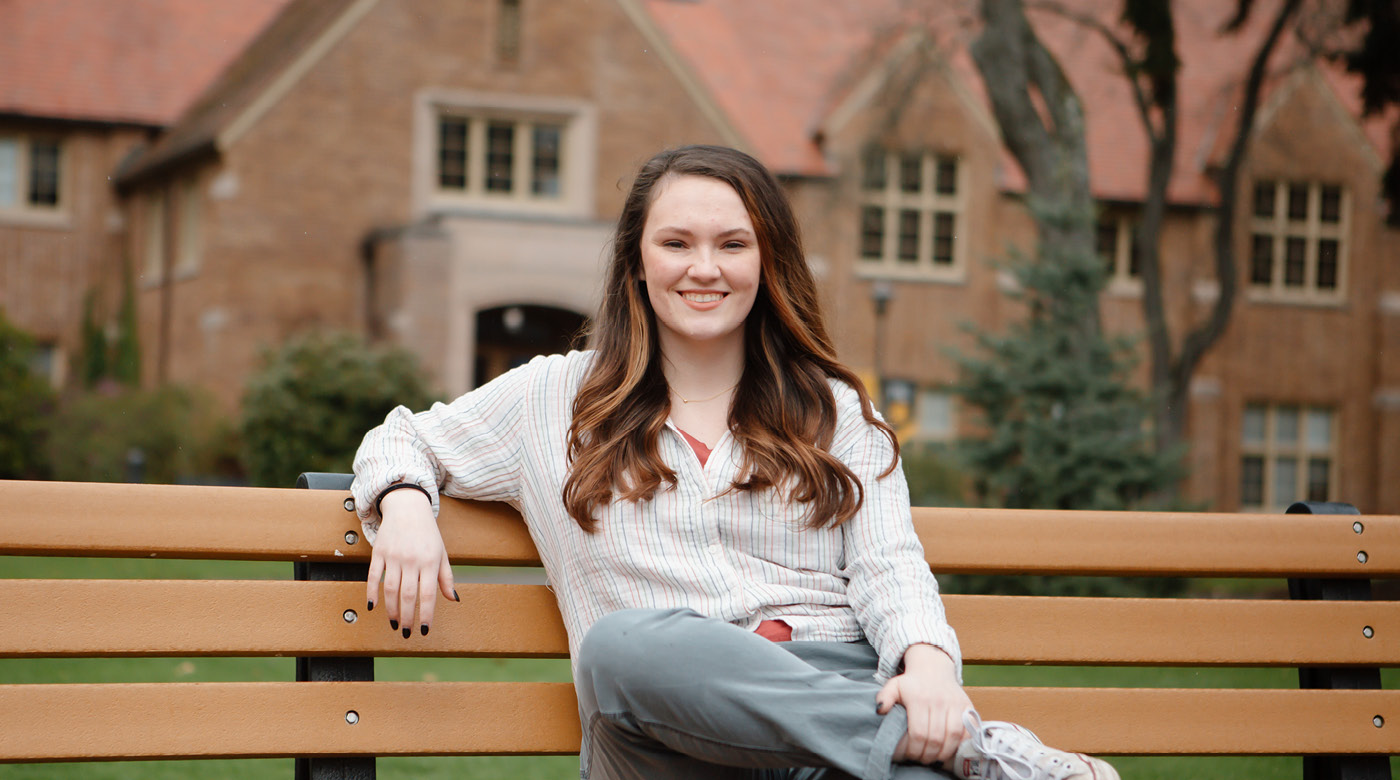 Kara Atkinson sits on a bench on PLU's upper campus with Xavier Hall behind her. She is smiling and has one of her arms wrapped around the drop of the bench.