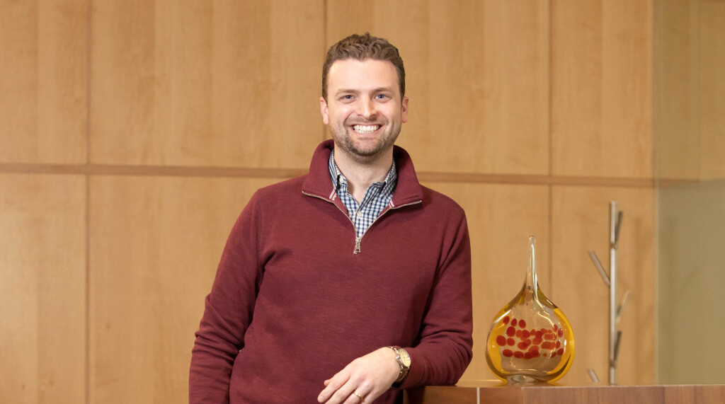 Michael Burris smiles in a conference room of his office. He is a learning against a high table and a piece of glass art sits next to him.