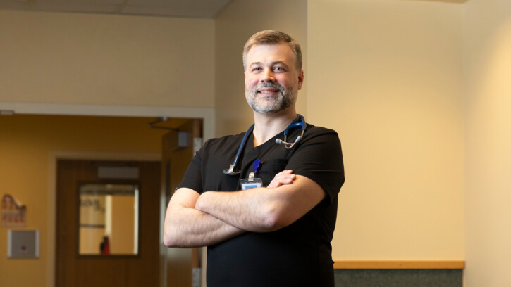 Dr. Erik Arnits '11 stands in a lobby of Samaritan Healthcare (hospital). He is wearing doctor scrubs, has his arms folded, and is smiling at the camera.